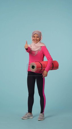 Photo for Full-sized isolated video capturing a young woman wearing a sportswear and a hijab, sheila. She holds an excercising mat and gives a thumbs up. Place for your advertisement, promotional, sport. - Royalty Free Image