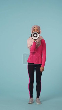 Photo for Full-sized isolated photo capturing an attractive young woman wearing a sportswear and a hijab, sheila. She is speaking through a rupor, loudspeaker. Place for your advertisement, promotional, sport. - Royalty Free Image