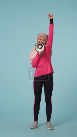 Foto de Foto aislada de tamaño completo que captura a una atractiva joven con ropa deportiva y un hiyab, Sheila. Ella está hablando a través de un rupor, altavoz. Lugar para tu anuncio, promoción, deporte. - Imagen libre de derechos