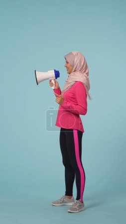 Photo for Full-sized isolated side view photo capturing an attractive young woman wearing a sportswear and a hijab, sheila. She is actively speaking in a rupor. Place for your adertisement, promotional, sport. - Royalty Free Image