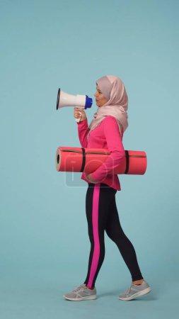Téléchargez les photos : Photo isolée pleine grandeur capturant une jeune femme portant un vêtement de sport et un hijab, Sheila, marchant à travers le cadre, parlant à travers un haut-parleur. Place pour votre publicité, promotion, sport - en image libre de droit