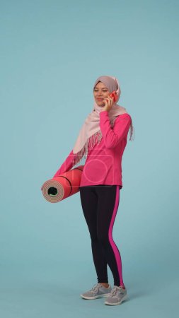Foto de Foto aislada de tamaño completo que captura a una joven con ropa deportiva y un hiyab, Sheila, sosteniendo una alfombra de ejercicio, hablando por teléfono. Lugar para tu anuncio, maqueta, promoción, deporte. - Imagen libre de derechos