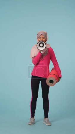 Photo for Full-sized isolated photo capturing an attractive young woman wearing a sportswear and a hijab, sheila. She is actively speaking in a loudspeaker. Place for your advertisement, promotional, sport. - Royalty Free Image