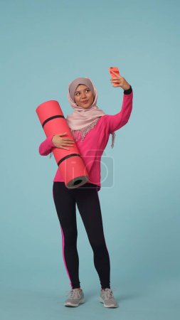 Photo for Full-sized isolated photo capturing a young woman wearing a sportswear and a hijab, sheila, holding an exercising mat, taking photos, selfies. Place for your advertisement, mock up, promotional, sport - Royalty Free Image