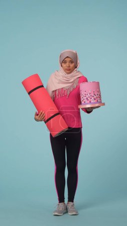 Photo for Full-sized isolated photo capturing a young woman in a sportswear and a hijab, sheila choosing between sport and junk food. Place for your advertisement, promotional, healthy lifestyle and sports. - Royalty Free Image