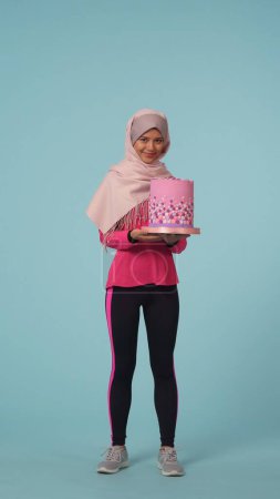 Photo for Full-sized isolated photo capturing a young woman in a sportswear and a hijab, sheila holding a cake, choosing junk food over healthy lifestyle. Place for your advertisement, promotional, sports. - Royalty Free Image