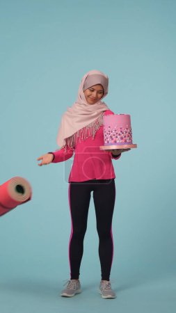 Téléchargez les photos : Photo isolée pleine grandeur capturant une jeune femme en tenue de sport et un hijab, Sheila choisissant le gâteau plutôt qu'un tapis d'exercice. Place pour votre publicité, promotion, mode de vie sain et sport. - en image libre de droit