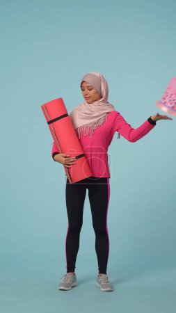 Téléchargez les photos : Photo isolée pleine grandeur capturant une jeune femme en tenue de sport et un hijab, Sheila choisissant le gâteau plutôt qu'un tapis d'exercice. Place pour votre publicité, promotion, mode de vie sain et sport. - en image libre de droit