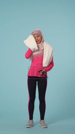 Téléchargez les photos : Photo isolée pleine grandeur capturant une jeune femme en tenue de sport et un hijab, Sheila tenant une corde à sauter après avoir fait de l'exercice et essuyé sa sueur sur le front. Place pour votre publicité, promo, sport - en image libre de droit