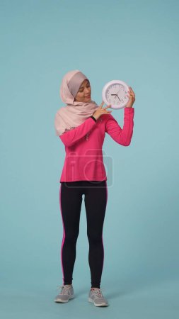 Téléchargez les photos : Photo isolée pleine grandeur capturant une jolie jeune femme portant un vêtement de sport et un hijab, Sheila. Elle tient une horloge et sourit. Place pour votre publicité, promotion, sport. - en image libre de droit