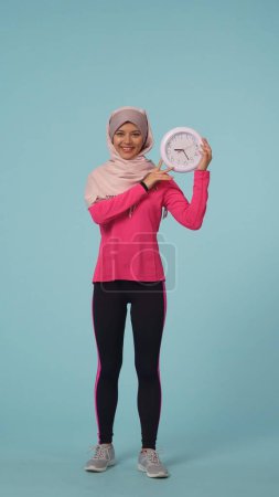 Foto de Foto aislada de tamaño completo que captura a una atractiva joven con ropa deportiva y un hiyab, Sheila. Ella sostiene un reloj y sonríe. Lugar para tu anuncio, promoción, deporte. - Imagen libre de derechos