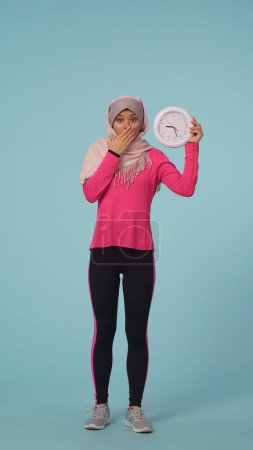 Foto de Foto aislada de tamaño completo que captura a una atractiva joven con ropa deportiva y un hiyab, Sheila. Ella sostiene un reloj, expresando confusión. Lugar para tu anuncio, promoción, deporte. - Imagen libre de derechos
