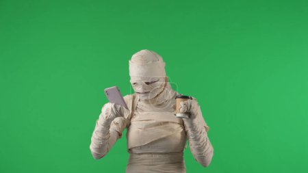Foto de Pantalla verde aislada croma clave foto capturar una momia celebración, escribiendo en un teléfono inteligente y sosteniendo una taza de café. Burla temática de Halloween, clip de promoción o anuncio de producto, ocio. - Imagen libre de derechos