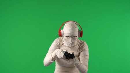 Photo for Green screen isolated chroma key photo capturing a mummy wearing headphones and holding a gamepad like its playing a videogame. Mock up, workspace for your promotion clip or advertisement. - Royalty Free Image