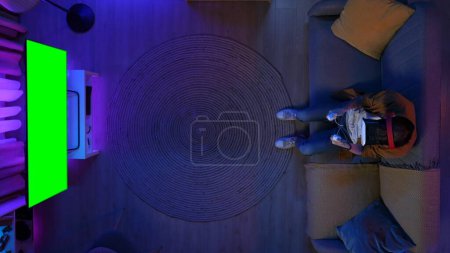 Foto de Concepto de publicidad de juegos y ocio. Vista superior del hombre sentado en el sofá en la habitación jugando juego en la consola de gafas VR, TV con pantalla verde tecla Chroma, espacio de trabajo área de publicidad maqueta. - Imagen libre de derechos
