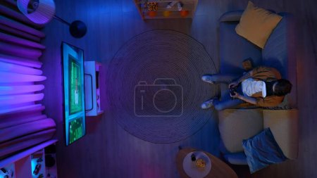 Photo for Online gaming and entertainment concept. Person spending free time at home. Top view of man sitting on the couch in the living room, wearing VR glasses playing action video game on TV . - Royalty Free Image