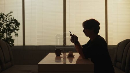 Photo for Everyday life and people relationships concept. Portrait of person in cafeteria. Silhouette of male sitting at the bar, holding smartphone and looking at the photos, dessert and coffee on the table. - Royalty Free Image
