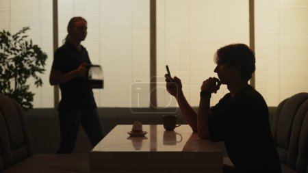 Photo for Everyday life and people relationships concept. Portrait of person in cafeteria. Silhouette of man sitting at the fast food bar holding reading social media on smartphone, waitress coming at the back. - Royalty Free Image