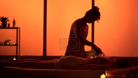 Foto de Masajista, masajista especialista en dar verter aceite esencial en la espalda del paciente. Siluetas en la sala de masajes, procedimiento de spa. Luces de neón exóticas y naranjas. Salud, terapia holística. - Imagen libre de derechos