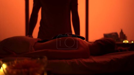 Téléchargez les photos : Masseur, spécialiste du massage offrant un massage relaxant en utilisant des roches chauffées. Silhouettes dans la salle de massage, procédure spa. Des néons exotiques, orange. Soins de santé, traitement médical, thérapie holistique. - en image libre de droit