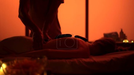 Photo for Masseur, massage specialist giving a relaxing massage using heated rocks. Silhouettes in the massaging room, spa procedure. Exotic, orange neon lights. Healthcare, medical treatment, holistic therapy. - Royalty Free Image