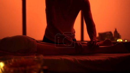 Foto de Un tiro medio. Masajista, especialista en masajes dando un masaje a su paciente. Siluetas en la sala de masajes, procedimiento de spa. Luces de neón exóticas y naranjas. Atención médica, tratamiento médico, terapia holística. - Imagen libre de derechos