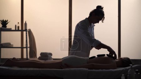 Téléchargez les photos : Masseur, spécialiste du massage offrant un massage relaxant en utilisant des roches chauffées. Silhouettes d'une femme et d'un homme dans la salle de massage, procédure spa. Soins de santé, traitement médical, thérapie holistique. - en image libre de droit