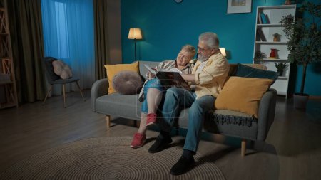 Foto de En el marco, una pareja de ancianos se sienta en un sofá en un apartamento, contra una pared azul. Están mirando, hojeando un álbum de fotos. Están charlando, recordando los viejos tiempos, nostálgicos. - Imagen libre de derechos