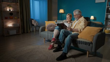Téléchargez les photos : Dans le cadre, un couple âgé est assis sur un canapé dans un appartement, contre un mur bleu. Ils regardent, feuilletant un album photo. Ils bavardent, se souviennent du bon vieux temps, nostalgiques. - en image libre de droit