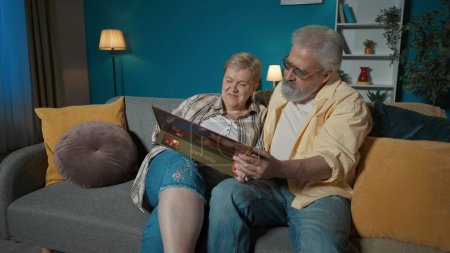 Téléchargez les photos : Dans le cadre, un couple âgé est assis sur un canapé dans un appartement, contre un mur bleu. Ils regardent un album photo. Ils bavardent, ils se souviennent du bon vieux temps, nostalgiques. Plan moyen. - en image libre de droit
