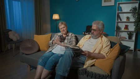 Téléchargez les photos : Sur la photo, le couple âgé s'assied sur le canapé de l'appartement contre le mur bleu. L'homme lit le livre à haute voix. La femme écoute, se réjouit et boit du thé. Confort à la maison. Plan moyen. - en image libre de droit