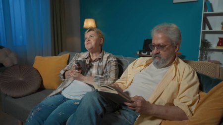 Foto de En la imagen de la pareja de ancianos se sientan en el sofá en el apartamento contra la pared azul. El hombre lee el libro en voz alta. La mujer escucha, se regocija y bebe té. Acogedor en casa. Tiro medio. - Imagen libre de derechos