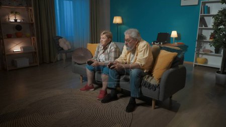 Foto de En la imagen, una pareja de ancianos está sentada en un sofá en una habitación. Están sosteniendo joysticks y mirando hacia otro lado. Demuestran el juego en la consola. Están atentos, enfocados. - Imagen libre de derechos