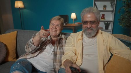 Téléchargez les photos : La vidéo montre un couple âgé assis sur un canapé dans une pièce contre un mur bleu. Ils détournent le regard comme s'ils regardaient une télévision. Ils sont heureux, riant. Plan moyen. - en image libre de droit