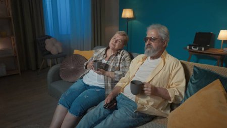 Téléchargez les photos : Sur la photo, une femme âgée est assise sur un canapé dans un appartement. L'homme lui a apporté du thé chaud. Ils regardent ensemble un téléfilm ou un programme. Ils sont heureux, souriants, prétendant aimer. - en image libre de droit