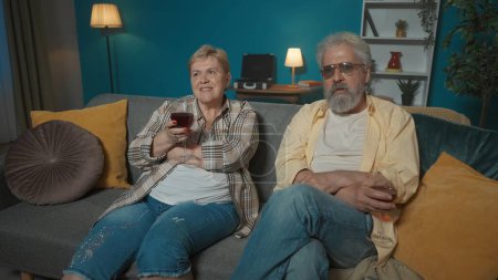 Photo for Frame shows an elderly couple sitting on a couch in a room. They have wine glasses in their hands, they are chatting pleasantly about something and drinking wine. They are happy, enjoying the evening. - Royalty Free Image
