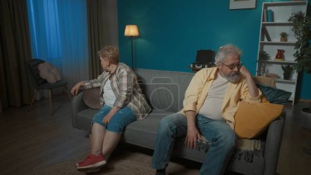 Foto de En el marco, una pareja de ancianos sentados en lados opuestos en un sofá. Demuestran pelea, resentimiento, ira, tristeza. Volviéndose el uno del otro, dicen algo, continúan peleando. - Imagen libre de derechos