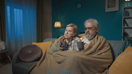 Foto de En la toma, una pareja de ancianos está sentada en un sofá debajo de una manta a cuadros con una taza de té o café. Están mirando a la distancia como si estuvieran en un televisor. Parecen serios, atentos.. - Imagen libre de derechos