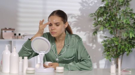 Photo for A young woman without makeup applying white cream on problem skin areas of the face. A woman in green silk pajamas sitting at a dressing table in front of a mirror in the morning. Problematic facial - Royalty Free Image
