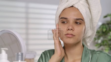 Foto de Retrato de una mujer joven después de una ducha frente a un espejo con una toalla de baño en la cabeza de cerca. Mujer examina su cara en el espejo para detectar arrugas e imperfecciones de la piel - Imagen libre de derechos