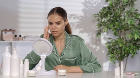 Téléchargez les photos : Portrait d'une femme frustrée à sa coiffeuse devant un miroir. La femme déçue souffre à cause de son problème de peau - en image libre de droit