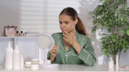 Photo for A young woman with problematic facial skin looks disappointedly in the mirror. A woman without makeup and wearing green silk pajamas sits at her dressing table in the morning. The concept of problem - Royalty Free Image