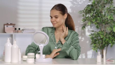 Photo for A woman without makeup and wearing silk pajamas, admires her reflection in the mirror while sitting at a dressing table. Problematic facial skin - Royalty Free Image