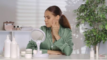 Photo for A woman doing her morning routine notices swelling, puffiness, or redness on her cheek. A young woman without makeup, wearing green silk pajamas sits at a dressing table in front of a mirror - Royalty Free Image