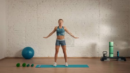 Photo for Personal sport classes at home online. Blonde female in sportswear doing exercises. Healthcare creative advertisement concept. Woman fitness coach in the room standing with hands wide open. - Royalty Free Image