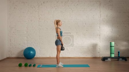 Photo for Personal sport classes at home online. Blonde female in sportswear doing exercises. Healthcare creative advertisement concept. Woman fitness coach in the room side view standing with kettlebell. - Royalty Free Image