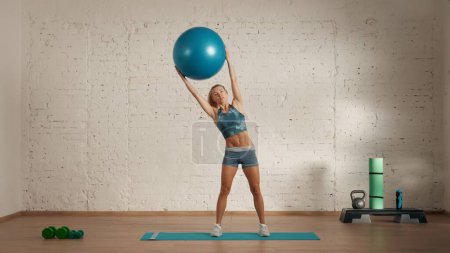 Photo for Personal sport classes at home online. Blonde female in sportswear doing exercises. Healthcare creative advertisement concept. Woman fitness coach in the room doing side tilts with fit ball. - Royalty Free Image