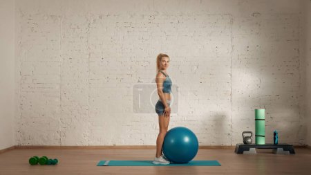 Photo for Personal sport classes at home online. Blonde female in sportswear doing exercises. Healthcare creative advertisement concept. Woman fitness coach in the room standing at the camera with ball. - Royalty Free Image