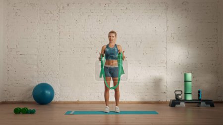 Photo for Personal sport classes at home online. Blonde female in sportswear doing exercises. Healthcare creative advertisement concept. Woman fitness coach in the room talking about rubber band. - Royalty Free Image