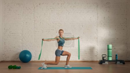 Photo for Personal sport classes at home online. Blonde female in sportswear doing exercises. Healthcare creative advertisement concept. Woman fitness coach in the room doing lunges with rubber band. - Royalty Free Image